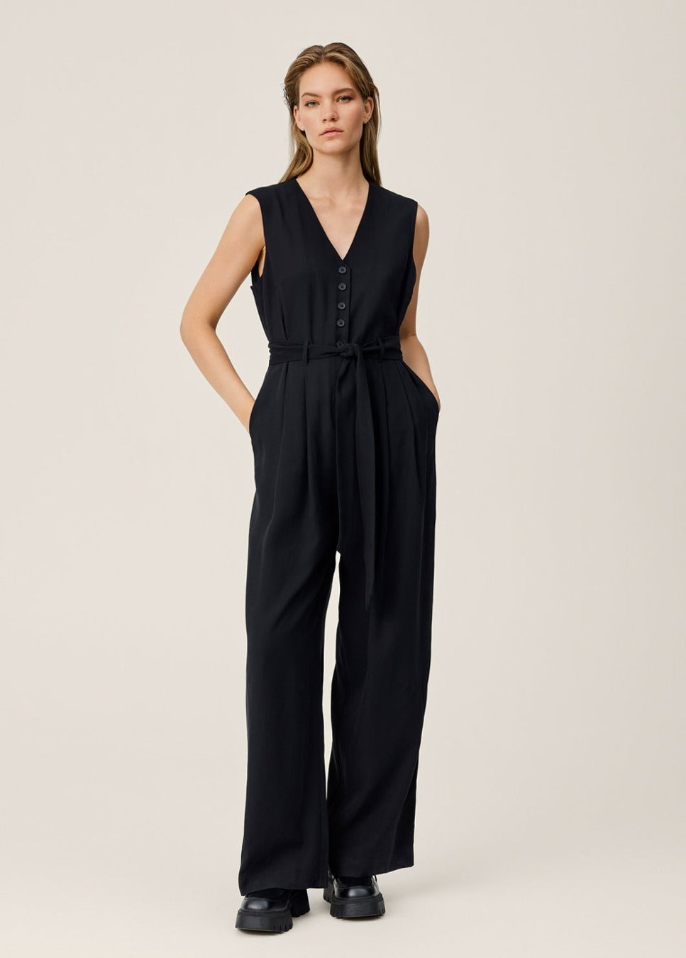 De zwarte mouwloze V-hals jumpsuit Asana van het merk MSCH Copenhagen. Deze zwarte, mouwloze jumpsuit heeft een elegante V-hals en is voorzien van een strikceintuur. De bovenkant van de jumpsuit heeft een sophisticated kostuumvest-achtige look met een knoopsluiting. Shop nieuwe jumpsuits bij dameskleding Boetiek Aniek.